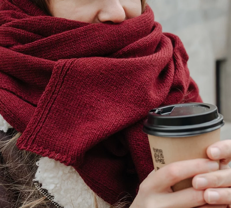 bundled up from the outside cold, woman stands wearing a scarf over her lower face holding a cup of coffee